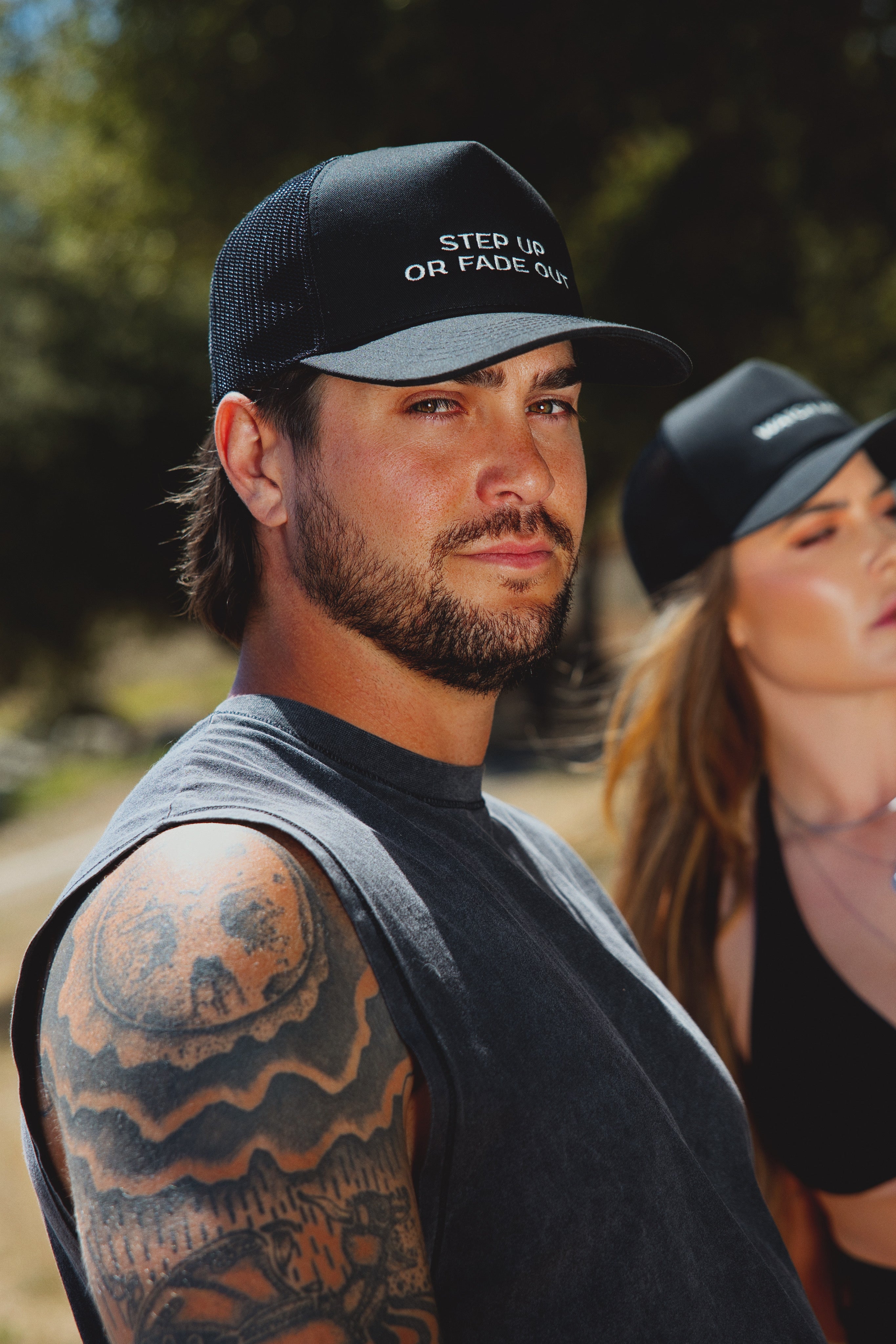 A man wearing a black trucker cap with white embroidered text reading 'STEP UP OR FADE OUT' in an outdoor setting, with a woman in a matching cap blurred in the background.