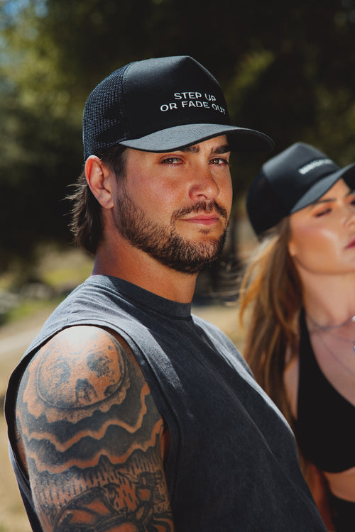 A man wearing a black trucker cap with white embroidered text reading 'STEP UP OR FADE OUT' in an outdoor setting, with a woman in a matching cap blurred in the background.